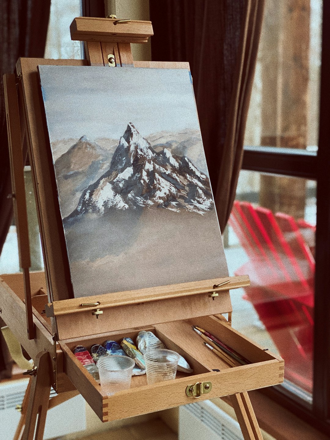 Acrylic mountain painting on an easel with scattered art supplies.