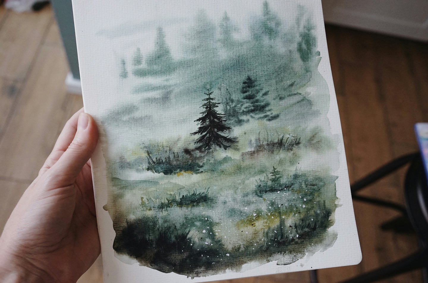 A hand holds a watercolor painting of a pine forest.