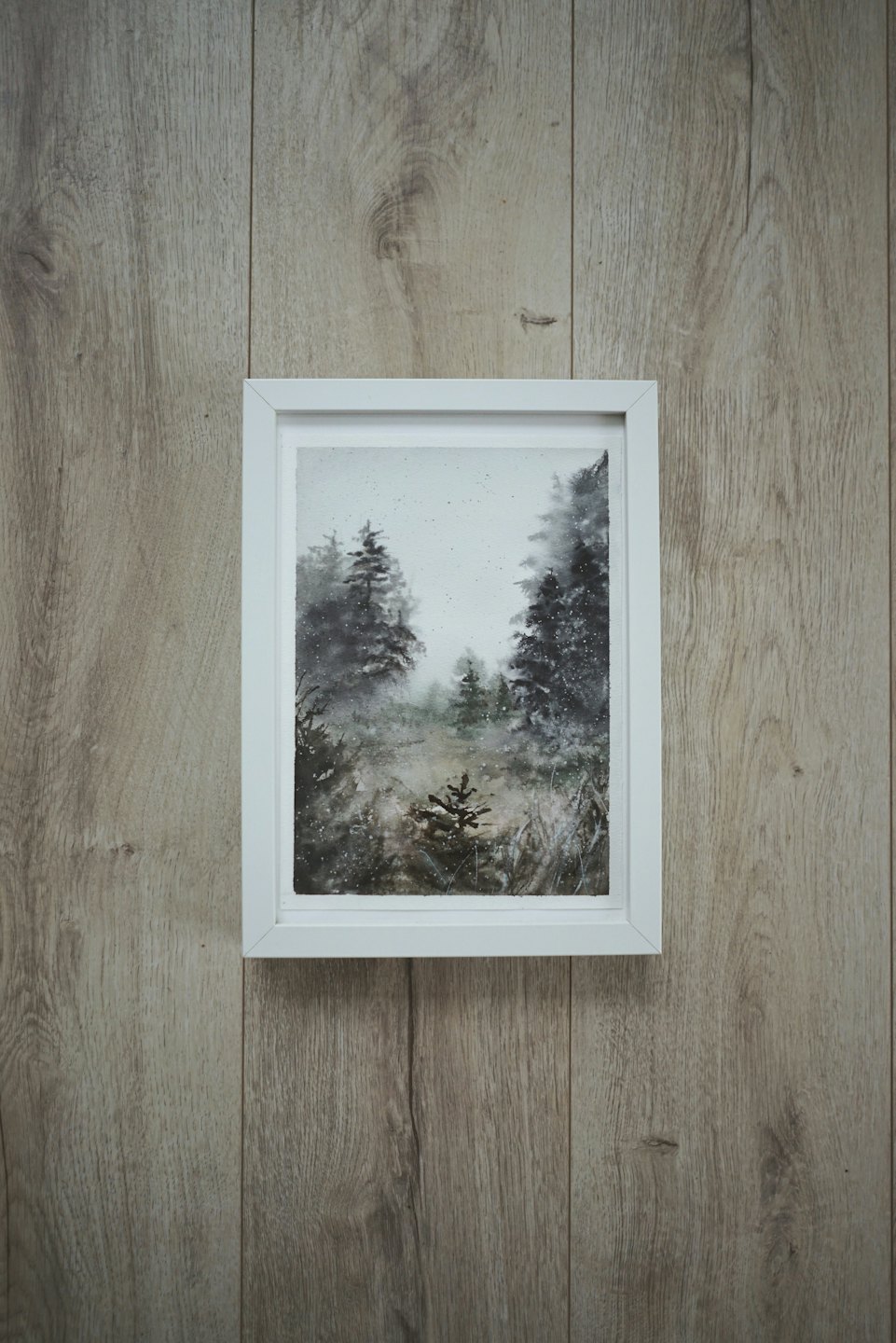 Framed watercolor painting of an evergreen forest on a wooden wall.